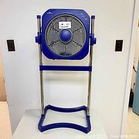 Front view of blue Air Innovations stand up circulating fan on metal and blue plastic stand showing fan blade and control panel.