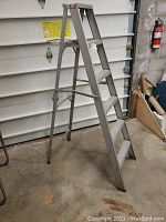Full view of 6-foot aluminum step ladder standing open on a concrete floor next to a garage door and fire extinguisher.