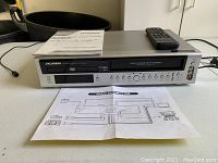 Sylvania VCR DVD combo unit displayed with remote control and instruction manual in front on a white surface.