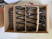 Four black coil suspension springs arranged in cardboard box compartments, showing overall condition and quantity.