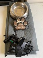 Photo showing all items: stainless steel dog bowl, black dog muzzle, grey car seat cover, and a paw-shaped decorative sign with text