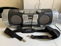 Front view of JVC boombox showing speakers, control panel, carrying strap laid in front with clips attached