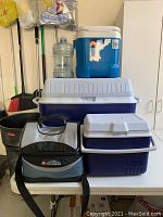 Photo displaying four coolers stacked or placed on a surface with brooms and other storage items in the background. Includes hard plastic coolers and one soft cooler bag along with a large clear plastic water bottle.