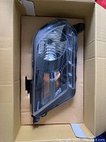 One headlight in original condition placed in cardboard box for protection. Black housing, clear lens, amber reflector and LED accents visible.