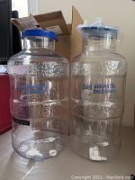 Two clear plastic Big Mouth Bubbler fermentors side by side with blue lids and white ported spigots at the bottom. The larger one has a temperature strip visible on the side.