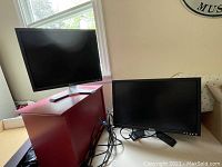 Two Dell computer monitors, one on a red box and another on the floor, showing size and style differences.