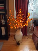 Tall ceramic vase holding an artificial flower arrangement with illuminated orange flowers. Plug-in cord visible at base.