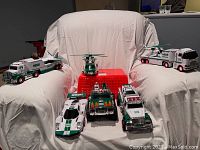 Photo showing multiple Hess trucks and vehicles arranged on a chair covered with white cloth including tanker truck, helicopter, semi-trucks, speedboat, racing car, and firetruck.