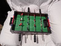 Angled side view of the tabletop foosball game on a white sheet showing the playing field, red and blue players, black handles, and one goal.