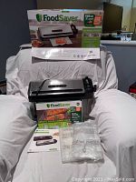 Photo showing Food Saver vacuum sealer machine placed on a white cloth with original box in background. Included are vacuum sealer bags and user manual.
