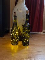Photo showing two green glass wine bottles each filled with warm yellow battery-powered fairy lights and sealed with cork stoppers on a wooden floor.