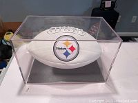 Front side of white collectible football showing Pittsburgh Steelers logo inside clear acrylic display case.