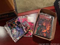 Photo showing a cardboard box filled with approximately 45 assorted comic books, some stacked outside the box, all in clear plastic sleeves.
