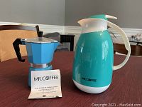 Photo of a turquoise Mr Coffee insulated coffee carafe and a blue aluminum 6 cup espresso maker with instruction booklet on a wooden table.