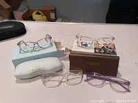 Four pairs of clear lens eyeglasses with clear frames, one with a purple tint. Two sets displayed on boxes, one in white protective case, one in brown YUN case, all set on a table.
