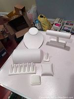 Photo showing various white jewelry display pieces including necklace bust, bracelet stands, ring holders, and pillows arranged on a white table with some background items like boxes and bags.