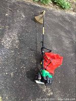 Full view of the Weed Eater showing the green engine, long shaft, black handle, and attached red accessory bag.