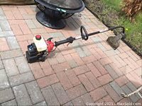 Side view of the full gas string trimmer showing the shaft, engine, fuel tank, and cutting guard on a brick patio.