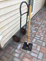 Photo showing the moving dolly and the tamper together against a beige siding wall on a paved area.