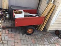 Full side view of red metal yard trailer with lumber and stakes inside, trailer box measures approximately 33x42x12 inches, showing considerable wear, one tire flat.