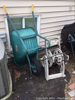 Wheelbarrow with teal resin body and metal frame standing against a wall next to a white plastic hose reel on ground with asphalt and some leaves.