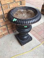 Front side of black cast iron urn with dirt, showing wear and paint peeling, placed against brick wall on concrete floor.