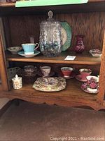 Two shelves of China cabinet displaying bone china teacups with saucers, small floral serving dishes, candy bowl, two small decorative vases, Royal Albert bowl, serving plate, and a clear glass candy jar with lid.