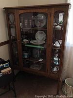 Full front view of the oak China cabinet showing glass front door, four shelves inside with some displayed china pieces, and side glass panels with carved wood detail.
