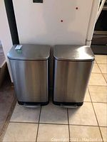 Two stainless steel garbage cans side by side on tiled floor, showing full front view.