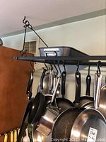 Photo showing black metal hanging pot rack mounted on ceiling with multiple pots and pans hanging on hooks along the bars.