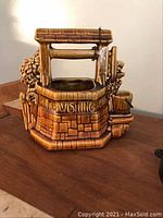 Front angle of brown ceramic wishing well cookie jar with textured stone and floral design, metal chain lid, and small bucket spout.