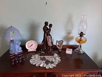 Full view of all items displayed on wooden furniture surface: Scottie lamp, oil lamp, Kiss statue, Big Ben clock, harp figurine, proverb cards, two small chairs, and a white clock.