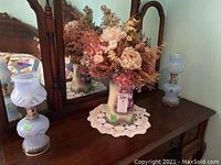 Pair of blue glass vintage lamps on a dresser with a large pitcher filled with faux flowers centered between them.