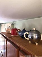 Four teapots placed in a row on a wooden shelf; includes the red, blue with metal cozy, white with red metal cozy, and cat-themed teapot.