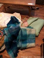 Four blankets and quilts displayed on floor and chair: two blue checkered Mohair throws, large green and tan wool blanket, and light-colored homemade quilt.