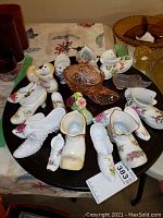 Top down view showing a variety of white porcelain shoes and boots with floral and gold detailing arranged on a table with a pink hobnail glass covered dish at center.