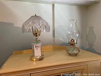 Electric lamp with patterned crystal base and large etched glass shade alongside a glass oil lamp with a round hobnail base and tall glass chimney shown together.
