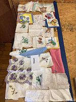 Full collection of vintage women's linen handkerchiefs spread out on a wooden surface, showing variety of designs including floral, animals, and people.