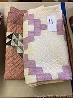 Two folded quilts. The top quilt has white background with lavender geometric pattern, the second quilt underneath shows black, white, and dusty pink pattern.