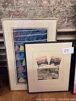 Two framed abstract watercolor paintings leaning against a brick wall. One larger, vividly colored with geometric design, the other showing three smaller earth-toned abstracts within a single frame.