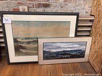 Two framed paintings leaning on a wooden floor against a brick and plywood wall. Left painting is a seascape in watercolor, right painting is a smaller landscape oil painting in greyscale.