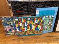 Three framed artworks leaning on floor: two colorful oil paintings and one silkscreen print in turquoise frame