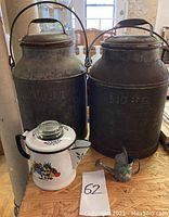 View of two large metal milk cans pressed with 'LiqBe' text, white tin enamel coffee pot with floral design, small decorated metal watering can - all placed on a wood surface.