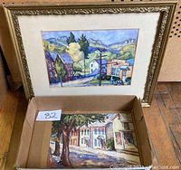 Framed watercolor painting alongside loose watercolor painting inside a cardboard box