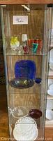 Full view of the glass display cabinet with all items arranged on three shelves; colors and varied glass shapes visible