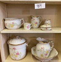Seven-piece ceramic chamber bath set arranged on shelves, showing washbowl, mug, chamber pots, soap dish, and toothbrush holder with floral patterns and gold trim.