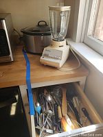 Photo of kitchen counter showing Farberware crock pot and Osterizer blender together with drawer open revealing kitchen utensils inside.