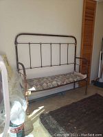 Front view of antique iron bench with quilted seat cushion, showing distressed rust and metal frame with vertical bars on backrest.