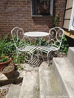 Full view of the metal patio table with two chairs outdoor against brick wall with plants.