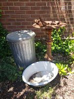 Iron bird bath, metal trash can, and enamel wash basin positioned outdoors against brick wall.